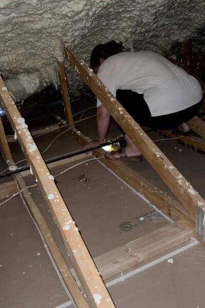 Crystel crouched between the attic trusses under spray foam insulation, installing a 2x4 between the truss bottoms so the room divider wall has something solid to attach to from below.