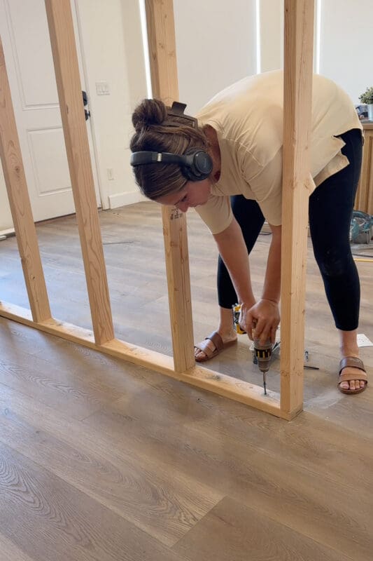 Crystel wearing ear protection and driving a self-tapping masonry screw through the bottom plate into the concrete slab where the flooring was cut away.