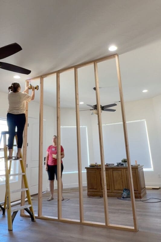 Crystel on a yellow ladder driving wood screws through the top plate into the ceiling blocking while her helper watches from below.