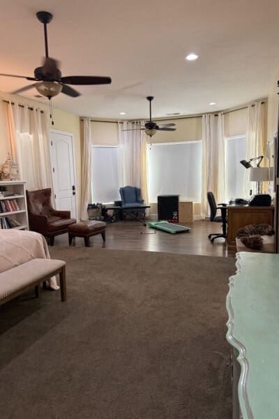 Long, open bedroom before the renovation, with carpet in the front sleeping area and wood flooring in the back, where a desk, weights, a walking pad, and a plyo box sit in front of the windows.