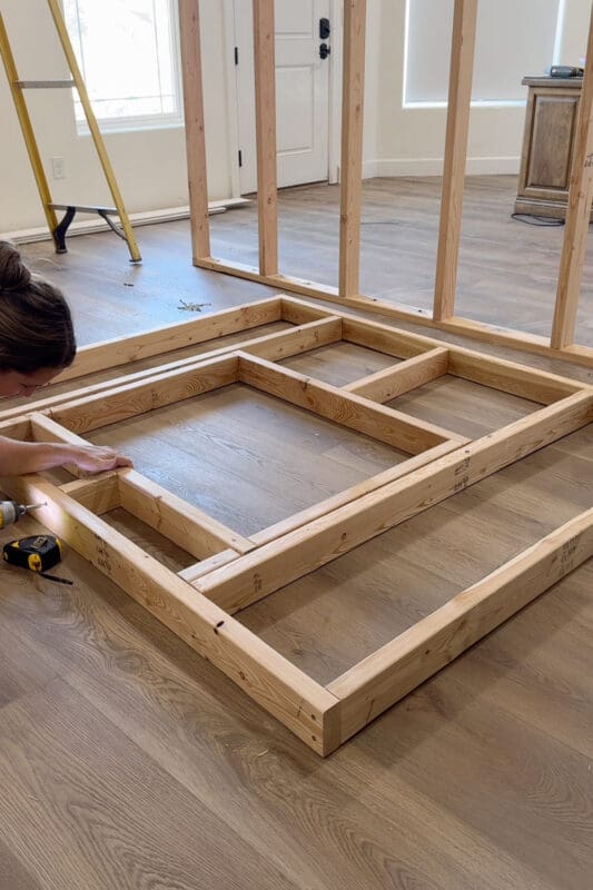 Crystel leaning over the fireplace insert frame on the floor, driving screws to join the inner and outer 2x4 boxes with cross braces, with a drill and tape measure beside her.