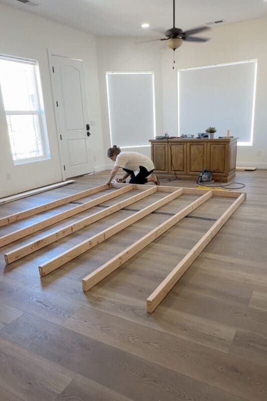 Crystel kneeling on the new plank flooring, screwing 2x4 studs to the top plate as she assembles the room divider wall frame flat on the floor.