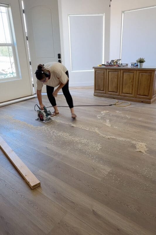 Crystel wearing ear protection and running a circular saw along the traced line in the plank flooring, with sawdust scattered across the floor and a wooden desk in the background.
