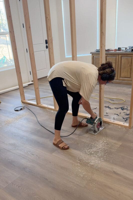 Crystel wearing ear protection and running a circular saw through the plank flooring in front of the framed room divider wall, with sawdust and screws scattered nearby.