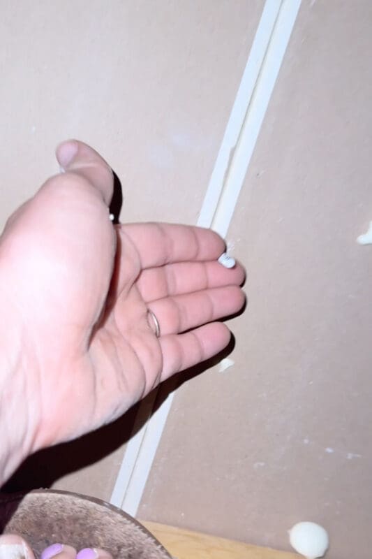 Crystel's hand pointing to a screw tip poking up through the back of the ceiling drywall in the attic, with spray foam drips on the surface around it.