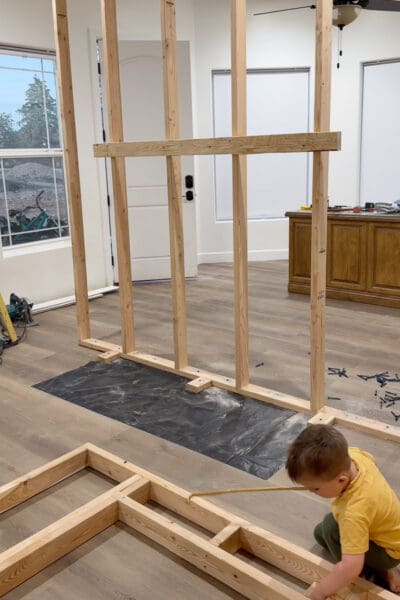 Room divider wall frame with a horizontal 2x4 across the studs and short 2x4 blocks set out from the bottom plate onto the moisture barrier, while a young helper plays with the fireplace insert frame in front.
