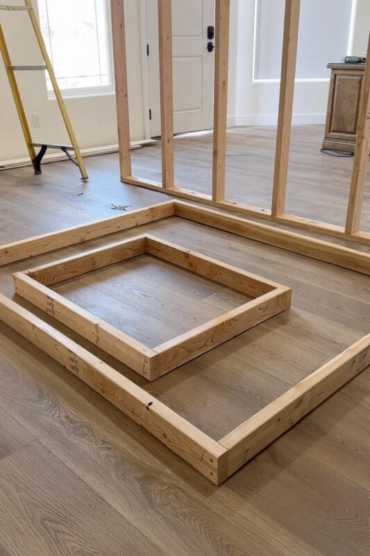 Two rectangular 2x4 frames laid flat on the plank flooring, one nested inside the other, as the framing for a fireplace insert takes shape in front of the standing room divider wall.
