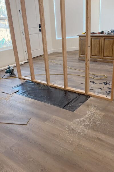 Rectangle of flooring removed in front of the stud frame down to the black moisture barrier, clearing space for the fireplace framing and a future hearth.