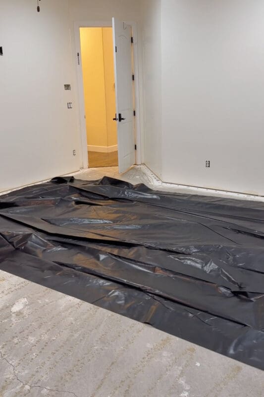 Bare concrete slab partly covered in a black plastic moisture barrier, with freshly painted white walls and an open door, as the flooring gets replaced before the room divider wall goes in.