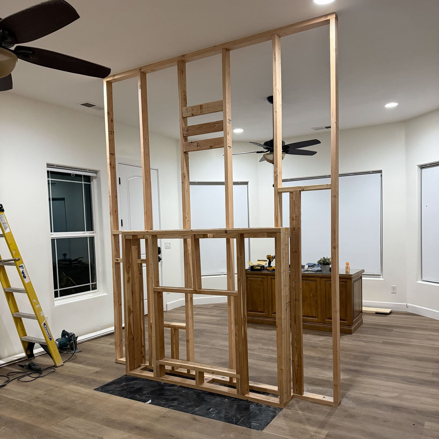 Freestanding wood stud frame for a DIY room divider wall in the middle of a large bedroom, with a lower framed opening for a fireplace insert, a yellow ladder, and a circular saw on the floor nearby.