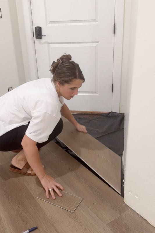 Crystel kneeling in front of a white door, fitting a wood-look plank into place over the black moisture barrier, with a cut piece of flooring and a marker on the finished floor beside her.