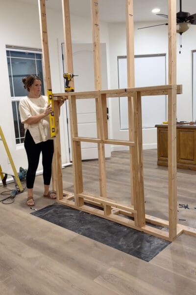 Crystel holding a yellow level against the fireplace insert frame, now attached to the bump-out blocking on the front of the room divider wall, with a drill resting on top of the frame.