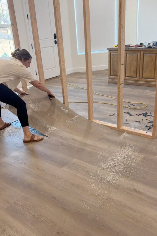 Crystel pulling up a section of cut plank flooring in front of the wall frame, revealing the black moisture barrier underneath.