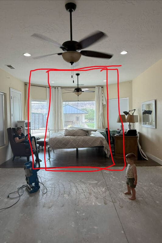 Large bedroom with the carpet pulled up to the concrete subfloor and a red hand-drawn outline showing where the room divider wall will go, with a woman sitting in a leather armchair and a young helper standing nearby.