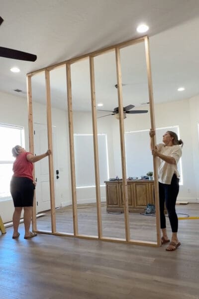 Crystel and a helper in a red shirt tilting the framed wall upright and guiding its top plate toward the ceiling.