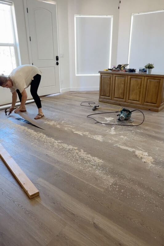Crystel using a hammer to pry up the cut strip of flooring, exposing the black moisture barrier underneath, with the circular saw resting on the floor nearby.