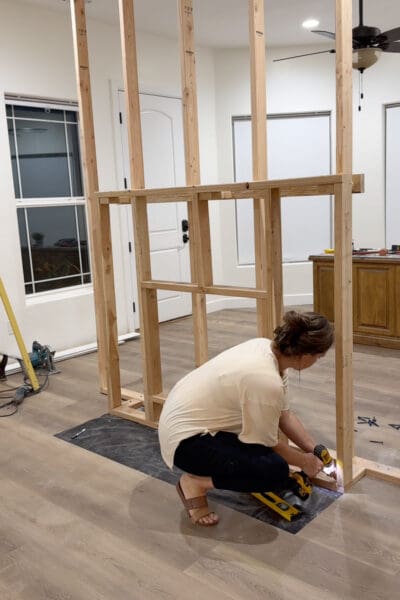 Crystel crouching beside the fireplace insert frame and driving a screw through its base into the bump-out blocking, with a yellow level on the moisture barrier beside her.
