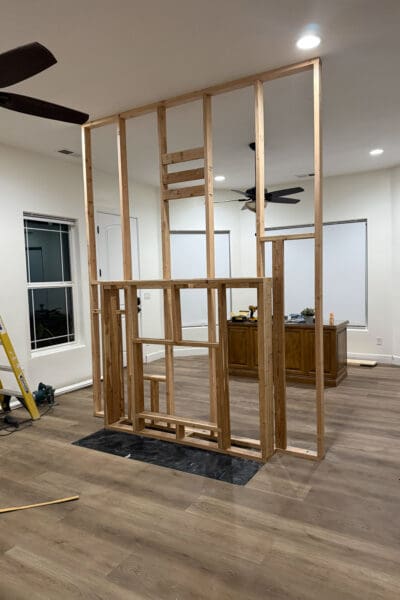 Alt text: Framed DIY room divider wall standing on new wood-look plank flooring, separating the bedroom from the back office area with its windows and a wooden desk holding a drill.