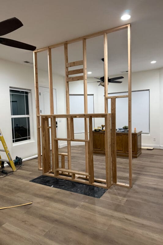 Alt text: Framed DIY room divider wall standing on new wood-look plank flooring, separating the bedroom from the back office area with its windows and a wooden desk holding a drill.