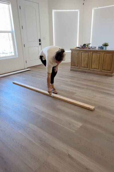 Crystel bending over a 2x4 laid on new wood-look plank flooring, tracing along its edge to mark where the room divider wall will stand.