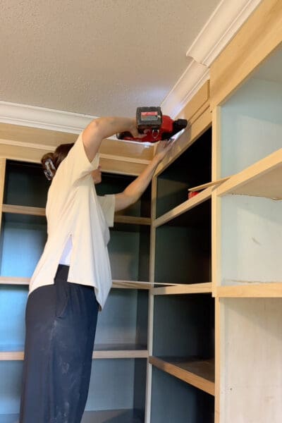 Crystel reaching overhead with a brad nailer to attach a strip of trim beneath the crown molding on the built-ins, using a spare board as a spacer for even placement.