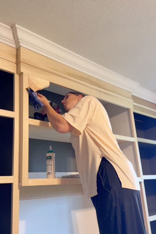 A blue clamp holds a curved wood corbel in place while Crystel drives a screw through it with a drill, creating the arched openings in the home library built-in bookshelves.