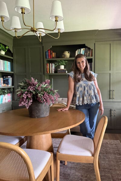 Crystel smiling beside the round table in her finished home library, with green built-in bookshelves and cabinets, aged brass pulls, a brass chandelier, and a dried floral arrangement in a stone pot.