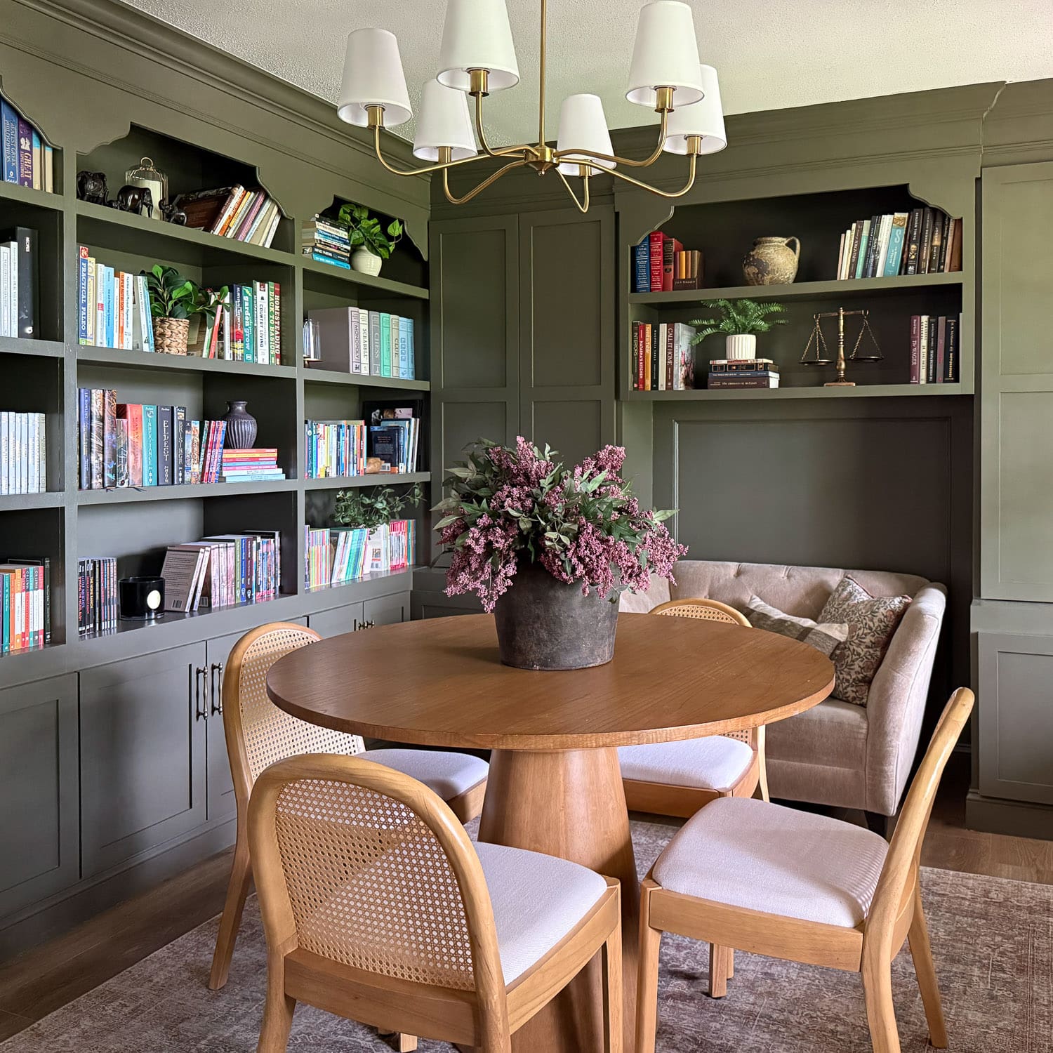 Finished home library makeover with walls of dark green built-in bookshelves, arched openings, brass chandelier, round wood table with cane chairs, and a tufted loveseat in the corner.