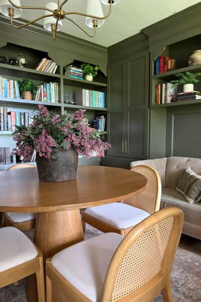 Corner view of the home library makeover showing painted green built-ins on two walls, arched shelf tops, paneled cabinet fronts, and a dried floral arrangement on the table.