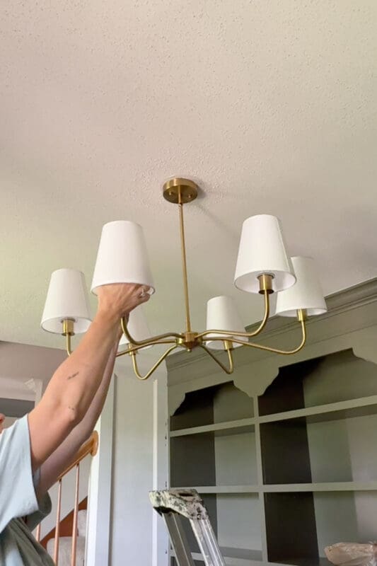Crystel steadying the new brass chandelier with white shades after hanging it in the home library, with the green built-in bookshelves and corbels in the background.