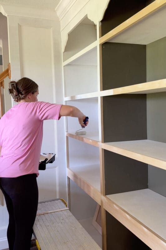 Crystel sitting on a work platform to reach the lower shelves, brushing primer onto the raw trim and shelf edges of the home library built-in bookshelves.