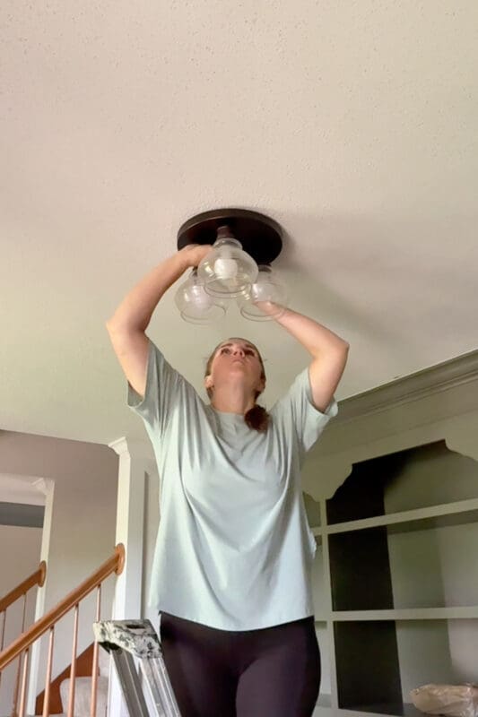 Crystel reaching overhead to unscrew the old three-globe flush mount light from the ceiling of the home library, with the painted green built-ins visible behind her.