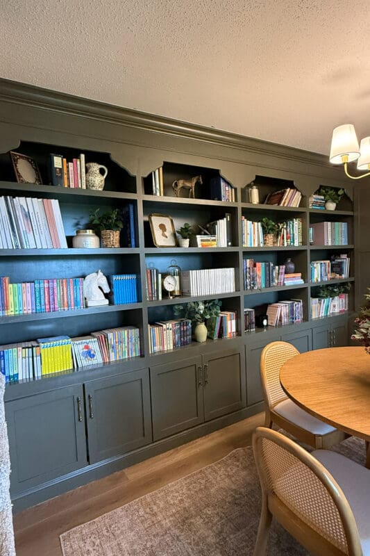 Full wall of green built-in bookshelves styled with color-sorted books, plants, framed art, and a brass horse figurine, with closed cabinet doors along the bottom.
