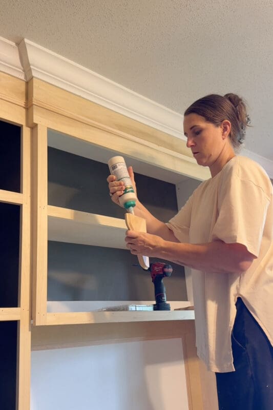 Crystel applying wood glue to the edge of a small curved corbel before attaching it to the corner of a bookshelf opening in the built-ins.