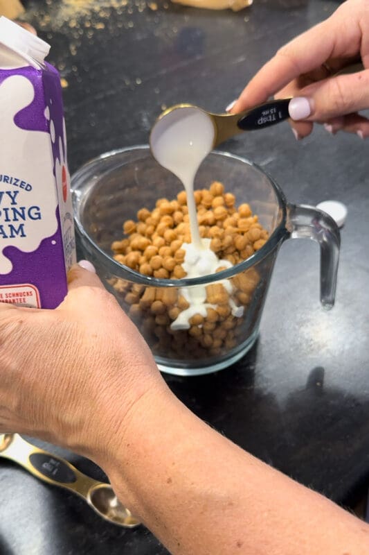 A hand pouring a tablespoon of heavy whipping cream over caramel bits in a glass measuring cup before microwaving