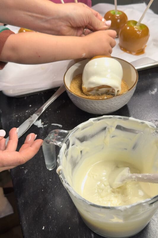 A young helper and an adult pressing a caramel apple coated in melted vanilla almond bark into a bowl of graham cracker crumbs, with caramel-dipped apples on a parchment-lined baking sheet behind them