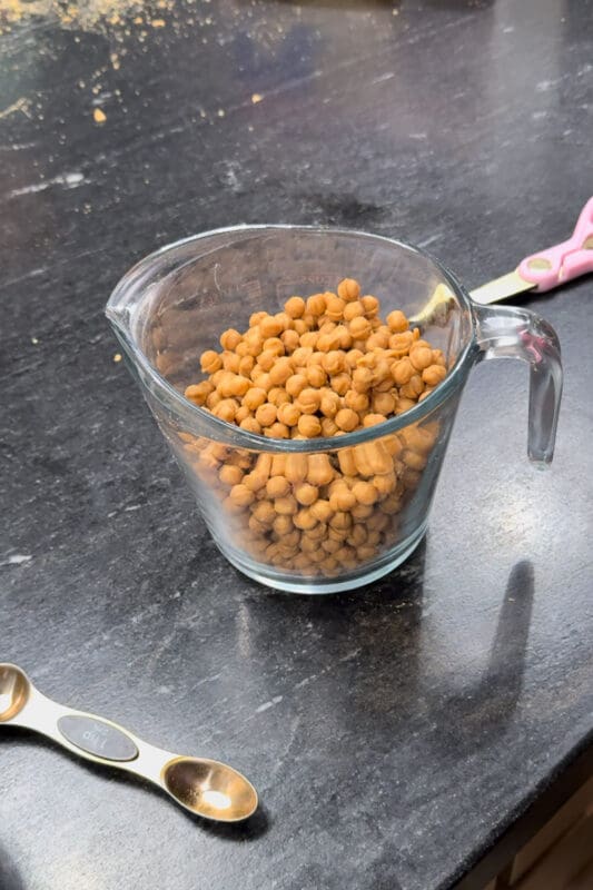 Caramel bits in a large glass measuring cup on a black kitchen counter, with gold measuring spoons nearby, ready to melt for apple pie caramel apples