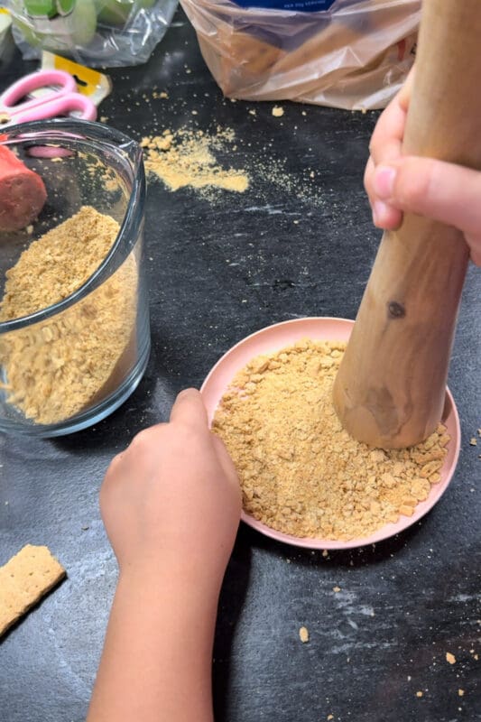 A young helper crushing graham crackers into fine crumbs with a wooden pestle on a small pink plate, with a glass measuring cup of crumbs nearby