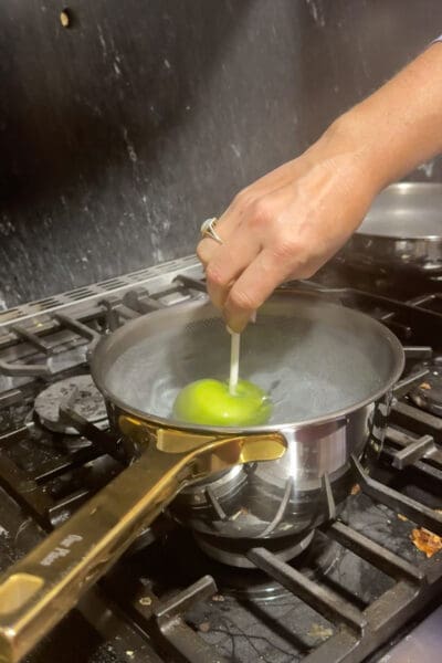 A hand dipping a green apple on a stick into a pot of steaming hot water on a gas stove to remove the wax coating so the caramel will stick
