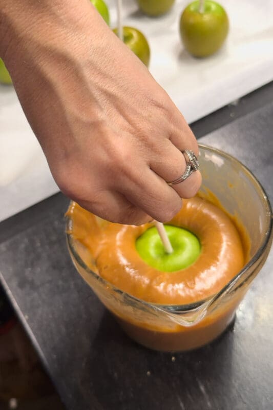 A hand dipping a green apple on a stick into a glass measuring cup of melted caramel, with more apples on sticks lined up on parchment paper in the background