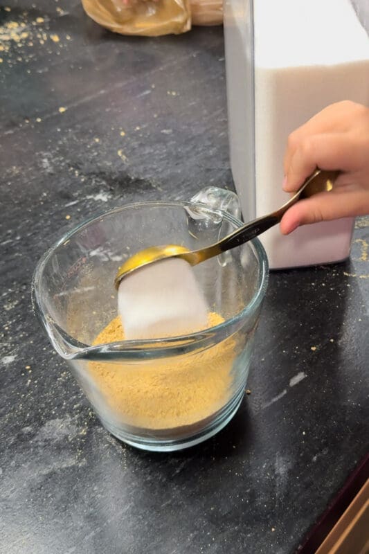A young helper adding a tablespoon of sugar to graham cracker crumbs in a glass measuring cup to make the apple pie topping