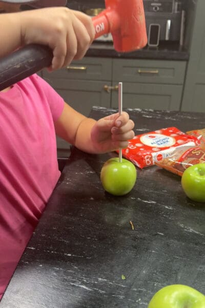 A young helper using a rubber mallet to tap a white stick into a green apple on a black kitchen counter, with a package of vanilla almond bark and more apples nearby, prepping for apple pie caramel apples