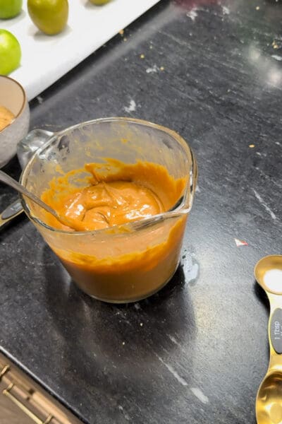 Smooth melted caramel stirred with a spoon in a glass measuring cup, with dewaxed green apples on sticks and a bowl of graham cracker crumbs waiting in the background