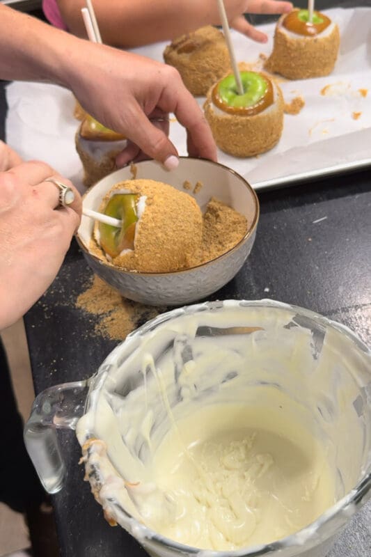 Hands rolling an apple pie caramel apple in a bowl of cinnamon sugar graham cracker crumbs, with finished apples lined up on parchment paper in the background