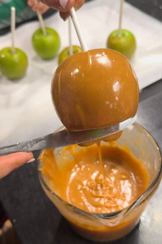 A caramel-coated apple held over the measuring cup while a butter knife scrapes excess caramel off the bottom, keeping the base of the caramel apple clean