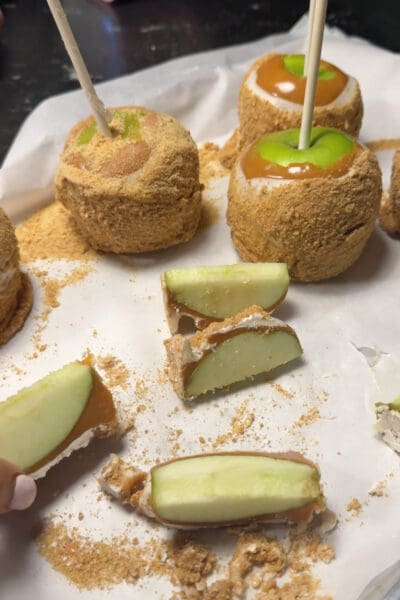 Sliced apple pie caramel apples on parchment paper showing the layers of green apple, caramel, vanilla almond bark, and graham cracker crumbs, with whole caramel apples on sticks behind them