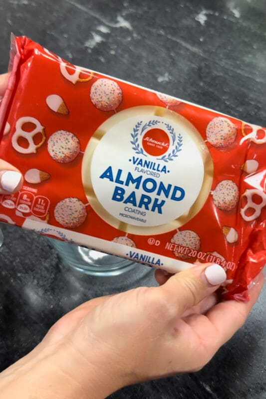 Hands holding a 20 oz package of vanilla almond bark coating over a glass measuring cup on a black kitchen counter