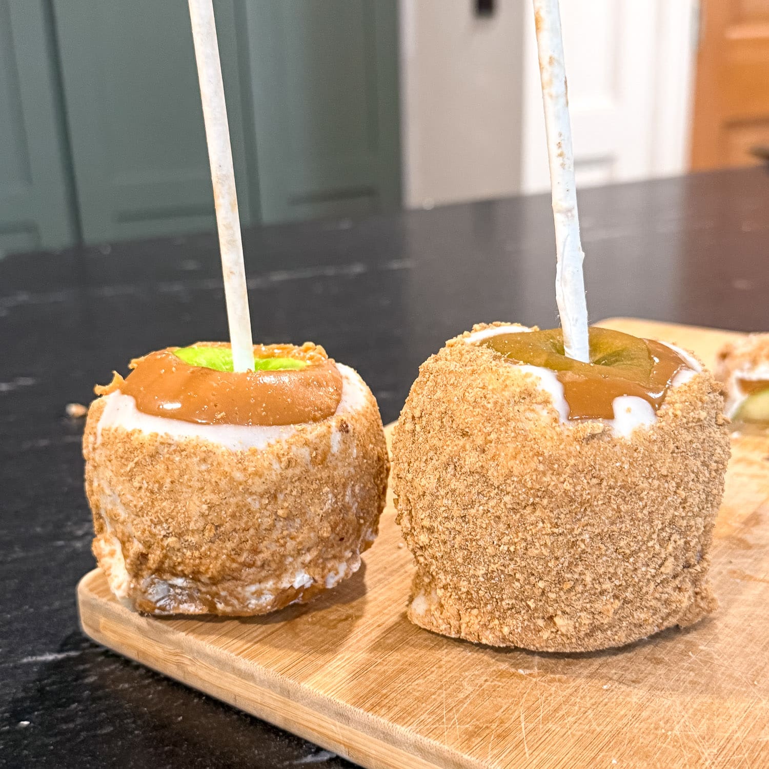 Two apple pie caramel apples on sticks coated in graham cracker crumbs, with caramel pooled on top and a layer of vanilla almond bark peeking through, sitting on a wooden cutting board in a kitchen with green cabinets