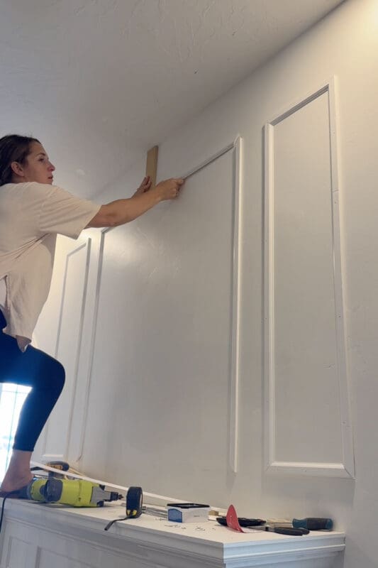 Crystel holding a scrap wood spacer against the ceiling to set the top rail of a new molding box, with finished picture frame molding boxes on the wall beside her for a large bedroom makeover