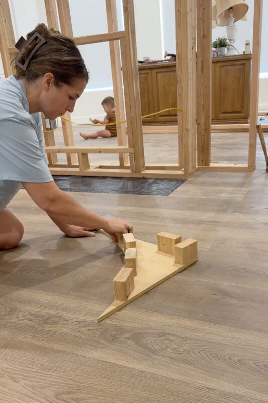 Crystel lining up short 2x4 blocks along a cut arch piece on the floor, with a young helper playing in the background behind the framed room divider wall